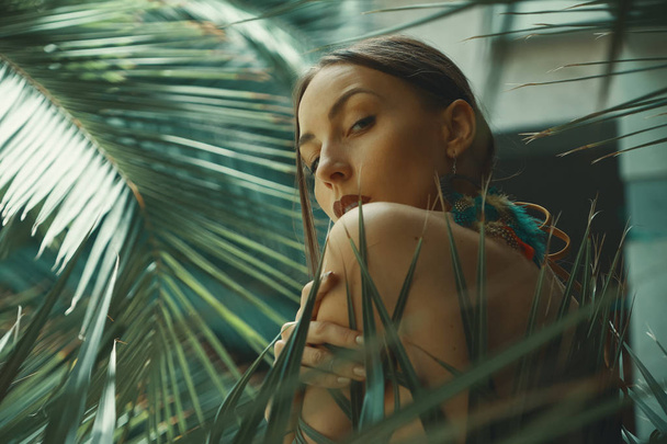 Retrato mulher exótica com ombros nus e brincos grandes com penas macias tocando com a mão sua pele enquanto relaxa entre as plantas tropicais. Copiar espaço à direita. Jogo de luz em folhas de palma
. - Foto, Imagem