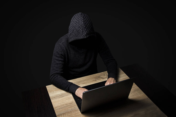 Faceless man in a hooded robe sits at a table with a laptop on a dark background. Concept of hacking and stealing user data. - Foto, Imagem