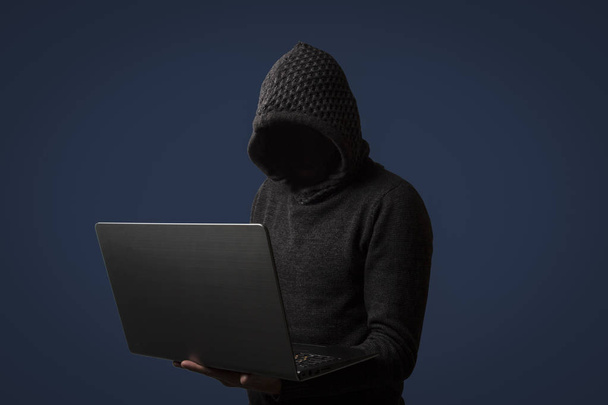 Faceless man in a hoodie with a hood holds a laptop in his hands on dark background. The concept of hacking and stealing user data. - Photo, Image