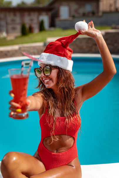 Jonge lachende vrouw in Kerstman hoed ontspannen in de buurt van zwemmen zwembad met glazen van cocktail - Foto, afbeelding