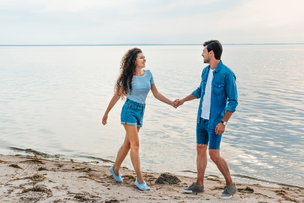 happy couple holding hands and walking on beach near sea  - Фото, изображение