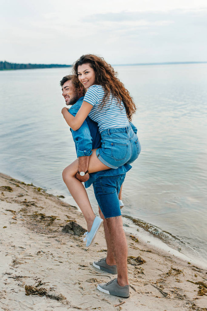 alegre joven pareja piggybacking en playa cerca de mar
  - Foto, Imagen