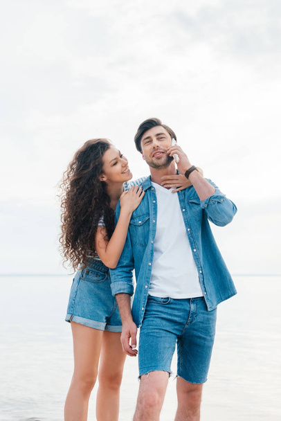 young girlfriend hugging her boyfriend while he talking on smartphone near sea   - Foto, Imagen