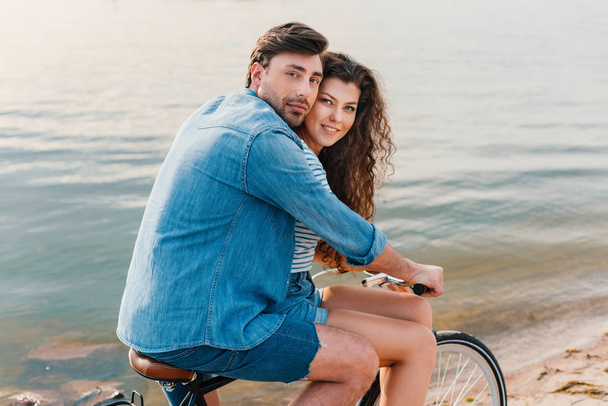 couple sitting on bicycle on beach near sea and looking at camera - Photo, image