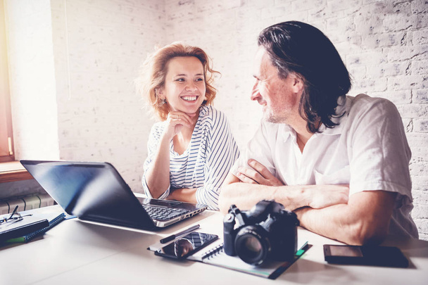 A middle-aged man and a young woman are sitting at a table with a laptop, camera and diary. Training and master classes in photography and processing, education concept, creative professions - Фото, изображение