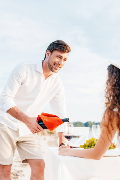 uśmiechnięty człowiek wylewanie czerwonego wina do kieliszka dla dziewczyny na romantyczną randkę na zewnątrz - Zdjęcie, obraz