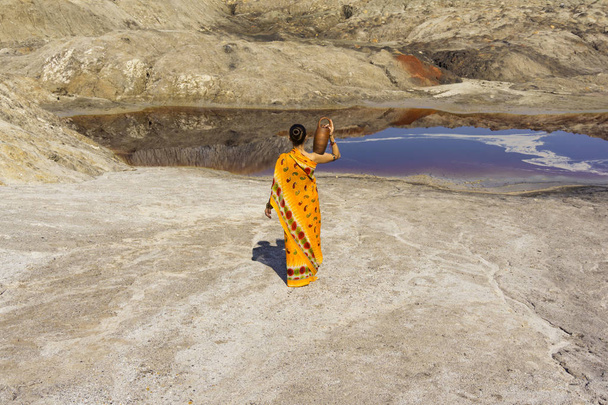 a girl in a sari with a pitcher on her shoulder goes for water to a dirty lake in a deserted landscape - Fotoğraf, Görsel
