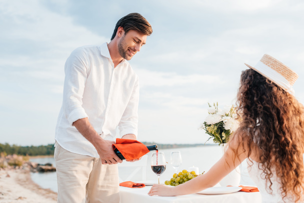 szczęśliwy człowiek wylewanie czerwonego wina do kieliszka dla kobiety na romantyczną randkę na plaży - Zdjęcie, obraz