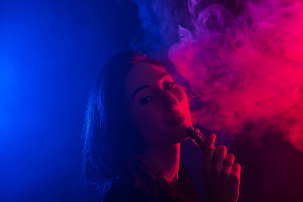 Портрет молодой женщины в неоновом красном и синем дыме с парой или электронными сигаретами
 - Фото, изображение