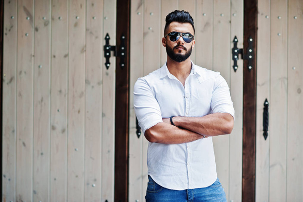 Stylish tall arabian man model in white shirt, jeans and sunglasses posed at street of city. Beard attractive arab guy. - Foto, immagini