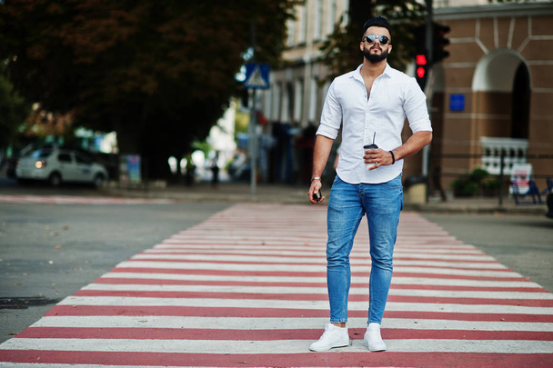 Stylish tall arabian man model in white shirt, jeans and sunglasses posed at street of city. Beard attractive arab guy with cup of coffee walking on pedestrian crossing or crosswalk. - Photo, Image