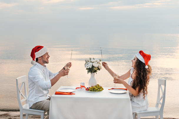 happy couple in santa hats celebrating christmas with champagne glasses and sparklers on beach - Foto, Imagem