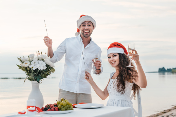couple in santa hats celebrating new year with champagne glasses and sparklers on beach - Foto, Imagem
