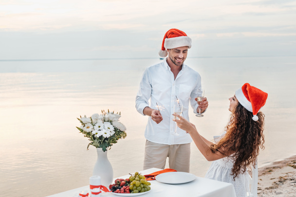 romantic couple in santa hats celebrating new year with champagne glasses and sparklers on beach - Photo, Image