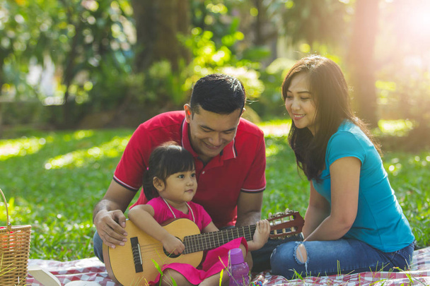 Папа и мама смотрят, как ее прекрасная дочь играет на гитаре в парке
 - Фото, изображение