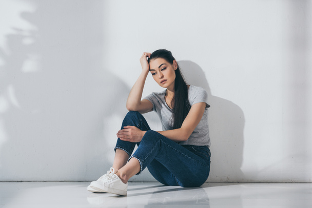 unhappy young brunette woman with hand on forehead sitting on floor near grey wall  - Foto, afbeelding
