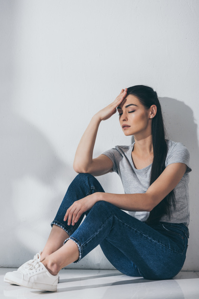 mujer joven infeliz con los ojos cerrados y la mano en la frente sentado en el suelo cerca de la pared gris
  - Foto, Imagen