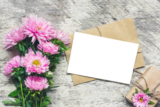 biglietto di auguri bianco vuoto con fiori rosa aster e scatola regalo su sfondo bianco in legno. posare piatto. Fai finta di niente. vista dall'alto
 - Foto, immagini