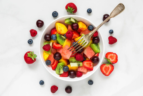 Obstsalat mit Wassermelone, Erdbeere, Kirsche, Blaubeere, Kiwi, Himbeere und Pfirsichen mit Gabel in einer Schüssel auf weißem Marmorhintergrund. Gesunde Ernährung. Ansicht von oben. - Foto, Bild