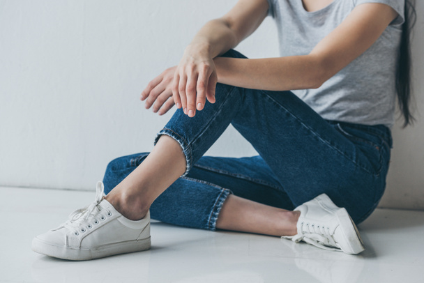 Schnappschuss eines Mädchens in Jeans, das auf dem Boden in der Nähe einer grauen Wand sitzt - Foto, Bild