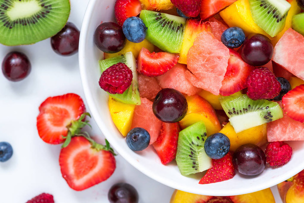 fruitsalade met watermeloen, aardbei, kersen, bosbessen, kiwi, framboos en perziken in een kom op witte marmeren achtergrond. gezonde voeding. Close-up. bovenaanzicht. - Foto, afbeelding