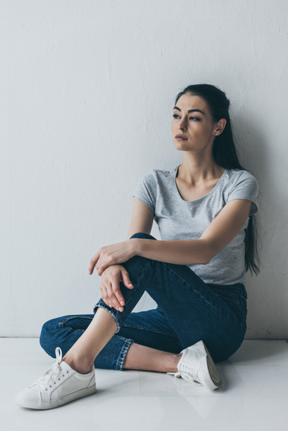depressive young brunette woman sitting near grey wall and looking away   - Foto, afbeelding