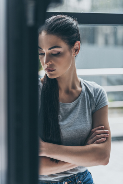 depresyjnych młoda brunetka kobiety stojącej z skrzyżowanymi rękami w pobliżu okna - Zdjęcie, obraz