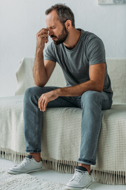 sad bearded man sitting on couch and rubbing nose bridge - Фото, изображение
