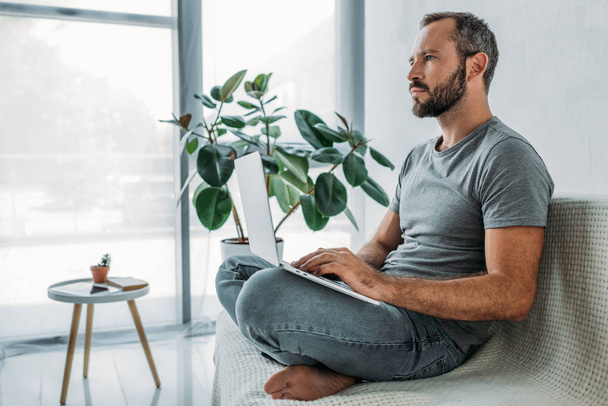 boční pohled zamyšlený vousatý muž sedí na gauči a pomocí přenosného počítače - Fotografie, Obrázek