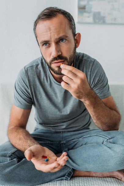 несчастный бородатый мужчина сидит на диване и принимает таблетки
 - Фото, изображение