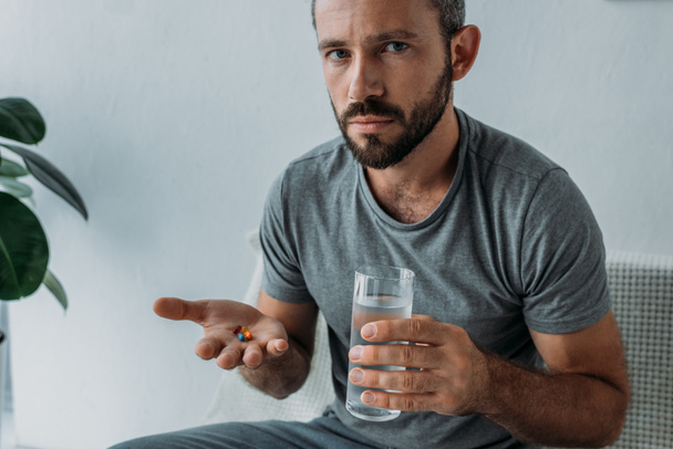 bearded middle aged man looking at camera while holding pills and glass of water - Photo, Image
