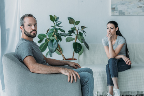 upset bearded man sitting on armchair and looking at camera while girlfriend sitting on couch behind, relationship difficulties concept   - Foto, Bild