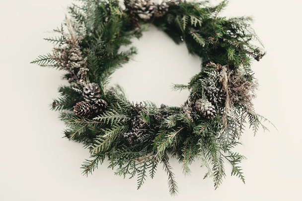 現代のクリスマスリース。松コーン、モミの枝、雪とスタイリッシュな素朴なクリスマスの花輪は、白い壁にかかっています。テキストのためのスペース。冬の休日のための手作りの装飾 - 写真・画像