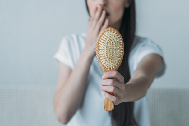 close-up view of shocked young woman holding hairbrush with fallen hair on grey  - Photo, Image