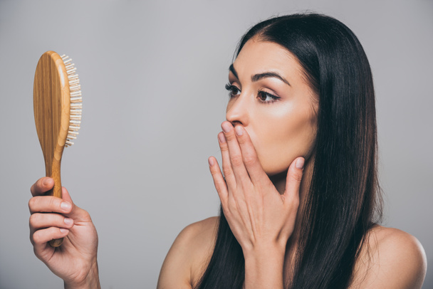 shocked young brunette woman looking at hairbrush isolated on grey, hair loss concept  - Φωτογραφία, εικόνα