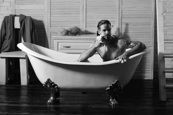 Macho sitting naked in bathtub washing with sponge. Man with beard and thoughtful face taking bath - Zdjęcie, obraz