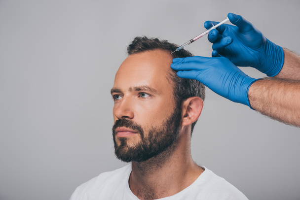 Schnappschuss eines Arztes mit Spritze, der einem Mann mit Alopezie eine Injektion verabreicht, der isoliert auf grau blickt   - Foto, Bild