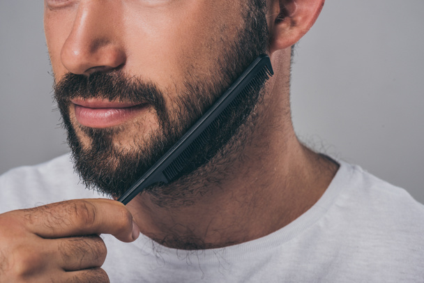 cropped shot of bearded man combing beard with comb isolated on grey - Photo, Image
