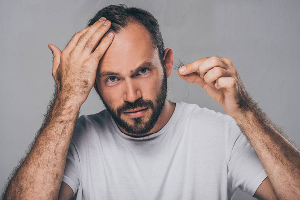 bearded middle aged man holding fallen hair and looking at camera isolated on grey - Photo, Image