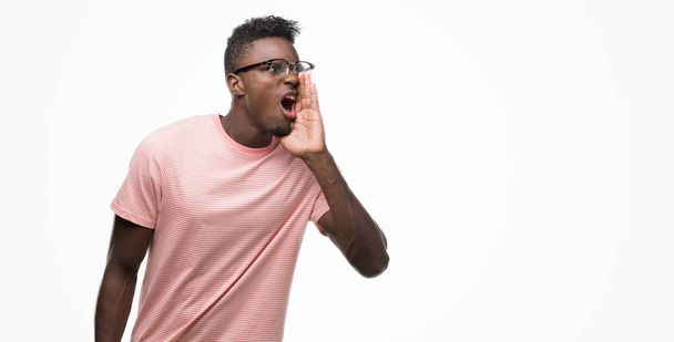 junger afrikanisch-amerikanischer Mann in rosa T-Shirt schreit und schreit laut mit der Hand auf dem Mund zur Seite. Kommunikationskonzept. - Foto, Bild