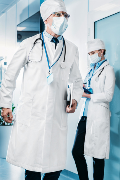 médecin masculin dans le masque médical marche avec tablette numérique tandis que sa collègue féminine debout derrière avec presse-papiers dans le couloir de l'hôpital
  - Photo, image