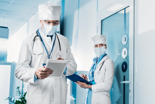 focused male doctor in medical mask using digital tablet while his female colleague standing behind with clipboard in hospital corridor  - Photo, Image