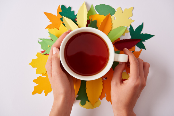 partial view of woman holding cup of hot tea on white tabletop with colorful papercrafted leaves - Photo, image