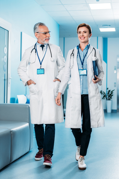 doctores sonrientes femeninos y masculinos con insignias y estetoscopios sobre el cuello caminando en el pasillo del hospital
  - Foto, imagen