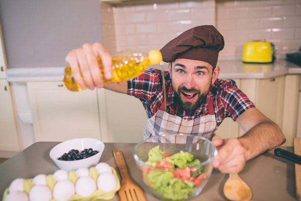 Amazed and happy guy stands at table in kitchen and holds bottle of oil. He poures some of it into bowl with salad. Guy keeps mouth opened. - 写真・画像