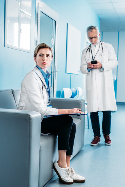 volwassen vrouwelijke arts op zoek weg en zit met Klembord op bank terwijl haar mannelijke collega gebruikend smartphone achter in de hal van het ziekenhuis - Foto, afbeelding