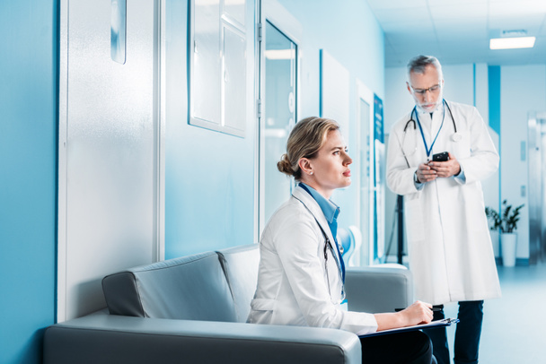 focused adult female doctor sitting with clipboard on sofa while her male colleague using smartphone behind in hospital corridor - Photo, Image