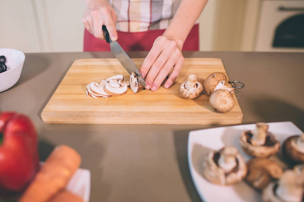Close up van vrouwtjes handen snijden paddenstoelen in stukjes. Ze doet dat voorzichtig. Meisje gebruikt mes. Er zijn plaat met solide champignons en andere groenten. - Foto, afbeelding