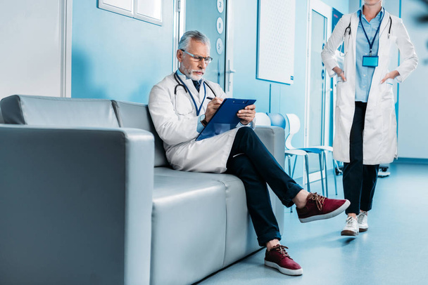 nadenkend midden leeftijd mannelijke arts schrijven in Klembord en zittend op de bank terwijl zijn vrouwelijke collega lopen achter in de hal van het ziekenhuis  - Foto, afbeelding