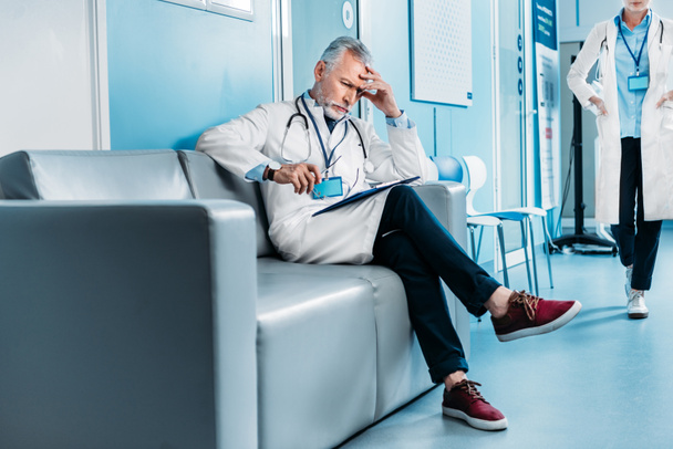 thoughtful middle aged male doctor with clipboard sitting on sofa while his female colleague walking behind in hospital corridor  - Photo, Image